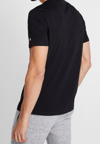 Black short-sleeve t-shirt with a round neck, made from smooth fabric; features a small logo on the left sleeve and is paired with gray pants.