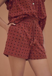 Shorts couleur rouille avec une taille à cordon de serrage, ornés de petits motifs géométriques noirs et d'accents à franges. Fabriqués en tissu léger et texturé.