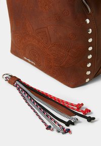 Borsa in pelle marrone con motivo floreale impresso, borchie argentate e una cerniera. Sei colorati pon-pon intrecciati con diversi dettagli nei nodi sono attaccati.