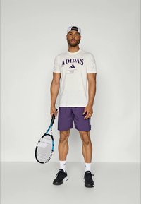 Hvit t-skjorte med kort erme og lilla logo, kombinert med lilla treningsshorts. Svarte joggesko og et tennisslag fullfører antrekket.