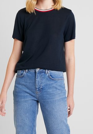 Granatowy T-shirt z krótkim rękawem, kołnierzyk w czerwono-białe paski, miękka tkanina, w zestawieniu z jasnoniebieskimi jeansami. Subtelny detal z logo przy krawędzi.