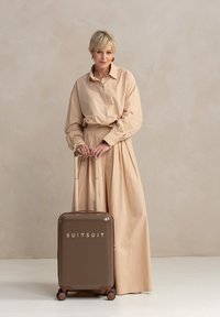 Een vrouw in een beige outfit staat naast een bruine koffer, gemarkeerd met "SUITSUIT," tegen een zachte, neutrale achtergrond.