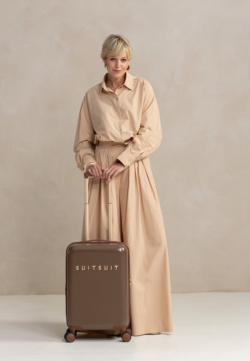 Een vrouw in een beige outfit staat naast een bruine koffer, gemarkeerd met "SUITSUIT," tegen een zachte, neutrale achtergrond.