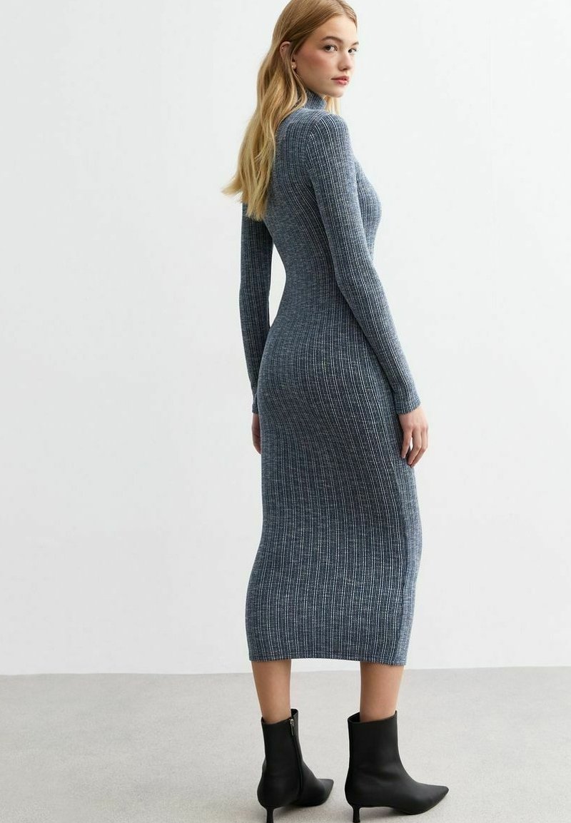 Abito lavorato a maglia di colore blu scuro con coste verticali, maniche lunghe, collo alto, silhouette aderente e lunghezza fino a metà polpaccio, abbinato a stivaletti neri alla caviglia.