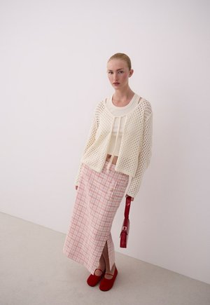 Frau trägt cremefarbenen, offen gestrickten Cardigan, weißes Top, rosa karierter langer Rock mit Schlitz, rote Schuhe und hält eine kleine rote Handtasche vor weißem Hintergrund.