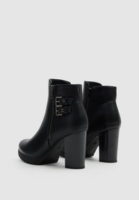 Botas de cuero negro hasta el tobillo con puntera redondeada, tacón alto y cuadrado, y cremallera lateral. Presentan detalles en forma de correas con hebillas de metal.