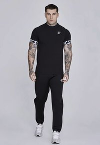 Melna īsroku krekls ar pelēku logotipu krūtīs, komplektā ar atbilstošām melnām biksēm. Nēsāts ar baltām sporta kurpēm. Gluda tekstūra.