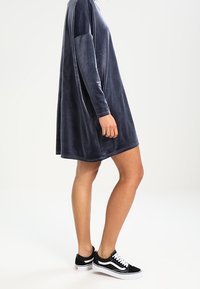 Sammet långärmad klänning i marinblå, lös passform, rundad fåll, kombinerad med svart-vita sneakers. Mjuk textur, minimal detaljering.