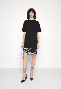 Czarna bawełniana sukienka t-shirtowa z krótkimi rękawami, z białym napisem "Karl" na dole, w zestawie z wielokolorową torebką i czarnymi szpilkami.