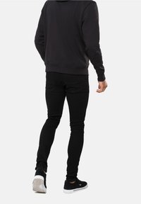 Una persona con una sudadera negra y jeans ajustados negros se aleja, vistiendo zapatillas negras, frente a un fondo blanco liso.
