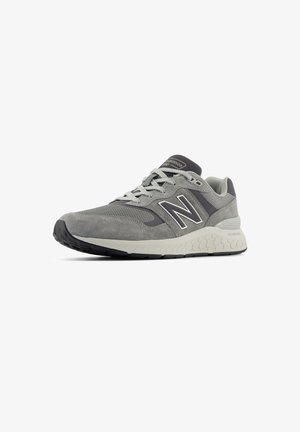 Zapatilla deportiva gris que presenta una combinación de materiales de ante y malla, con una suela texturizada y un detalle de logo reflectante.