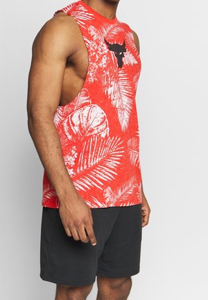 Persona musculosa llevando una camiseta sin mangas roja con hojas tropicales blancas y un logo de toro negro, combinada con pantalones cortos negros.
