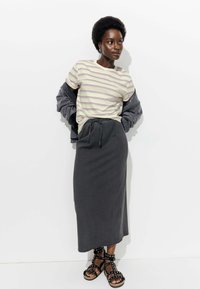 T-shirt rayé en crème, lavande et vert clair, associé à une jupe maxi en coton grise et des sandales noires à clous. Veste en jean drapée sur les bras.