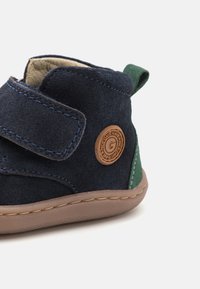 Bota de camurça azul-escura com detalhe verde, tira de velcro, forro bege, emblema redondo e sola de borracha castanha. Projetada para bebés.