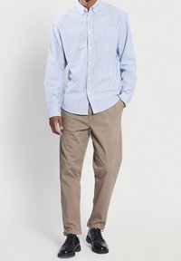 Camicia blu chiaro a righe con maniche lunghe, abbinata a pantaloni beige e scarpe nere. Presenta una tasca frontale e un colletto a fascia.
