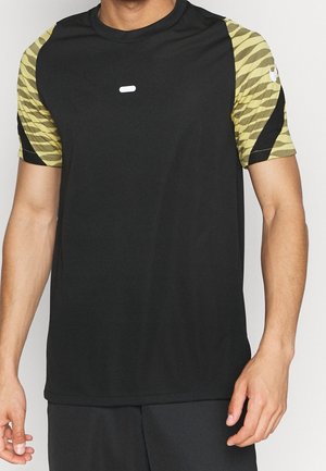 Mann trägt ein schwarzes Sportshirt mit gelb-schwarz gemusterten kurzen Ärmeln, steht vor weißem Hintergrund.