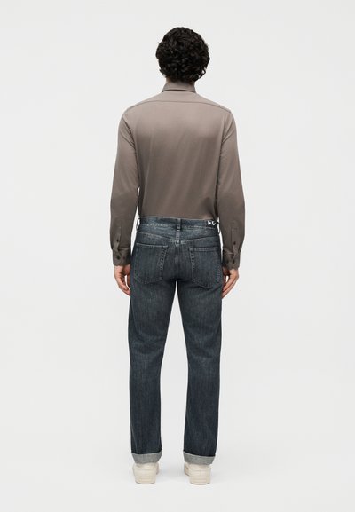 Άνδρας με σγουρά μαλλιά στέκεται με την πλάτη γυρισμένη, φοράει μπλούζα μακρυμάνικη σε απόχρωση ταμπά, σκουρόχρωμα τζιν με ρεβέρ και λευκά αθλητικά παπούτσια, σε απλό φόντο.