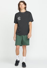 Zwarte korte mouwen t-shirt met een grafisch ontwerp op de linkerkant van de borst, gecombineerd met groene shorts, zwarte sneakers en witte sokken.