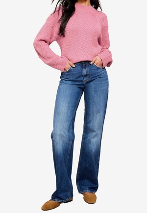 Femme portant un pull en maille texturée rose, un jean bleu taille haute à jambes larges et des chaussures plates couleur camel, mains dans les poches.