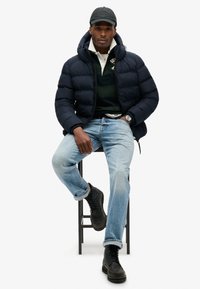 Marine pufferjakke, grøn og hvid polo, lyseblå jeans, sorte snørebåndsstøvler. Designet med hætte og logo-detaljer, siddende på en skammel.