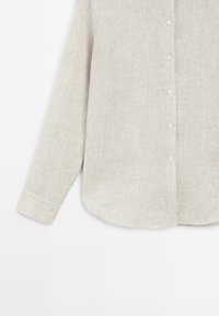 Jasno beżowa koszula zapinana na guziki o strukturze prążkowanej, z długimi rękawami i zaokrąglonym dołem. Posiada małe guziki z przodu.