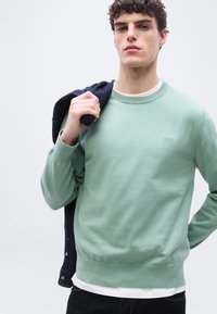 Jeune homme portant un pull vert menthe par-dessus une chemise blanche, tenant une veste foncée sur son épaule, debout devant un fond uni.