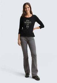 Black long-sleeve T-shirt with a round neckline featuring gold text "the season of joy," paired with gray flared jeans and black footwear.