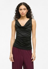 Top senza maniche nero con scollatura a cascata, dettagli drappeggiati sui lati, realizzato in tessuto morbido, abbinato a pantaloni bordeaux a pieghe.