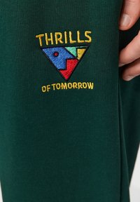 Zelená látka s vyšitým trojúhelníkovým logem v modré, červené, žluté a zelené barvě, a textem "THRILLS OF TOMORROW" ve žluté.