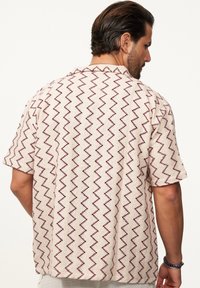 Kurzarmhemd aus beigem Stoff mit rotem Zickzackmuster, lockere Passform und Kragen. Strukturiert mit feiner Webart.