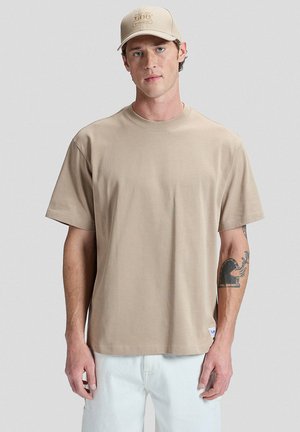 Mies, joka pitää beigeä lippalakkia ja vastaavan väristä ylipintaista t-paitaa, valkoiset housut, vasemmassa kyynärvarressa tatuointi, seisoo yksinkertaisen vaalean taustan edessä.