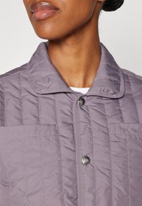 Prošívaná světle fialová bunda s plochým límcem, stříbrným zapínáním na knoflíky a dvěma náprsními kapsami. Materiál vypadá měkký a texturovaný.