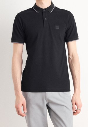 Man draagt een zwart poloshirt met korte mouwen met grijze bies op de kraag en mouwen, gecombineerd met lichtgrijze broek.