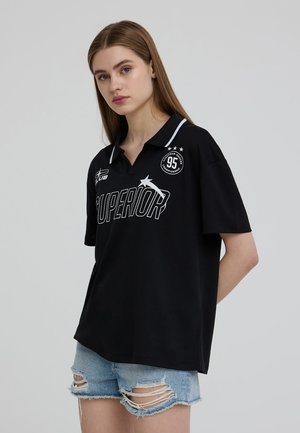 Polo nero con accenti bianchi e la scritta "SUPERIOR" stampata in modo prominente. Presenta un colletto, maniche corte e una vestibilità casual. Shorts in denim strappato.