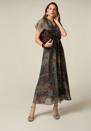 Vrouw die een lange paisley jurk draagt en een bruine gevlochten clutch vasthoudt, geaccessoriseerd met grote oorbellen, een armband en teal kleurige open-toe hakken.