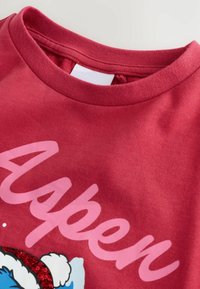 Sweatshirt in cotone rosso con scollatura a girocollo, testo rosa che recita "Aspen" e una grafica con un personaggio in cartone animato e decorazioni festive.