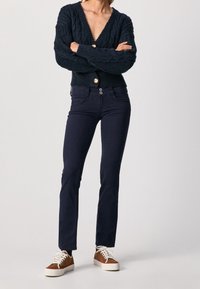 Marinblå kabelstickad cardigan med djup V-ringning och gyllene knappar, kombinerad med slim-fit marinblå jeans och bruna mockaskor.