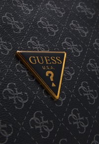 Materiale nero texturizzato con un motivo grigio ripetuto. Logo triangolare dorato con la scritta "GUESS U.S.A." e un punto interrogativo al centro.