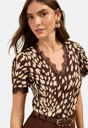 Frau trägt eine braune und beige Bluse mit Tiermuster und Spitzenbesatz, goldene, strukturierte Ohrringe und braune Hosen mit einem strukturierten Gürtel.