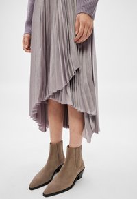 Moteris dėvi levandų spalvos klostuotą sijoną su asimetrišku kraštu ir taupės spalvos zomšines kulkšnies aulinukus, stovinčią ant balto fono.