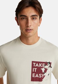 Mladý muž v béžovém tričku s grafikou Goofyho, který dává palec nahoru, a textem "TAKE IT EASY" v bordovém čtverci.