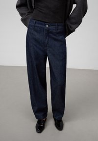 Mörkblå denimbyxor med avslappnad passform, knappstängning, framfickor och uppvikta benslut, kompletterade med svarta eleganta skor.