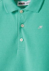 Grünes Polohemd aus strukturiertem Stoff, mit drei Knöpfen und einem gestickten "M"-Logo in Hellrosa auf der linken Seite.
