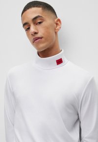 Polo de manga larga blanca con cuello alto que presenta una etiqueta roja de "HUGO". Textura suave, diseño ajustado y estilo minimalista.