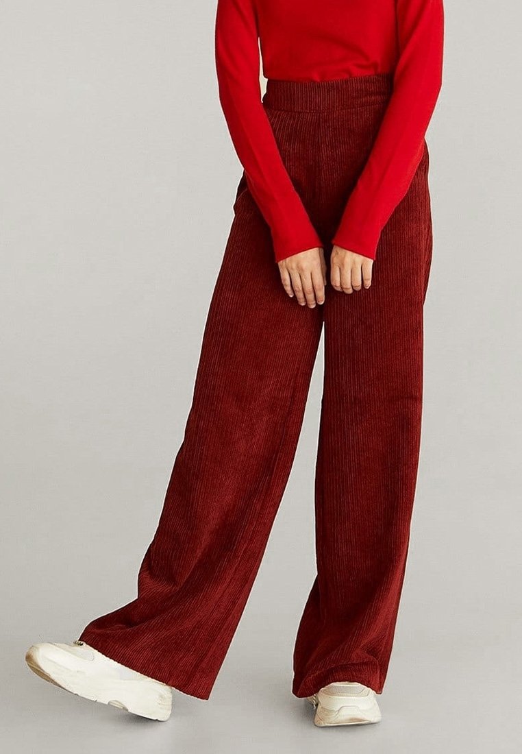 Pantalons en velours côtelé à jambes larges de couleur bordeaux avec des nervures texturées, associés à un haut rouge à manches longues et des baskets blanches.