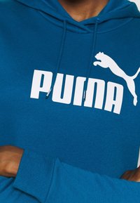 Modrá mikina s kapucí vyrobená z měkkého materiálu, s velkým bílým logem "PUMA" a grafikou skáčící pumy. Nastavitelná šňůrka v kapuci.