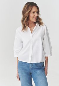 Camicia bianca con bottoni realizzata in tessuto leggero, caratterizzata da una vestibilità rilassata, maniche a tre quarti e colletto. Indossata con jeans blu.