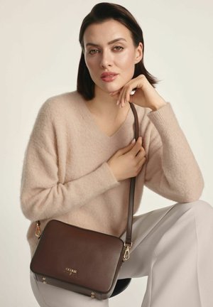 Kobieta z krótkimi ciemnymi włosami, ubrana w beżowy sweter i jasne spodnie, siedząca z ciemno brązową skórzaną torbą na biodro z złotymi akcentami.