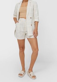 Blazer en tartan beige clair avec boutons marron, associé à un débardeur côtelé beige et un short tailleur assorti. Porté avec des sandales légères.