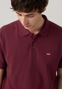 Burgundi rövid ujjú pólóing texturált anyagból, három gombos gombolással és Levi's logós folttal a bal mellen.